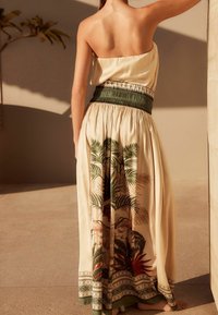 Vestido maxi con diseño sin tirantes, color crema, cinturilla de acento verde y estampado tropical con hojas de palma y detalles florales.