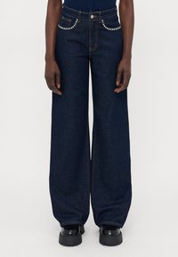 Vide leg mørke denimjeans med høy midje, med naglede detaljer på lommene og oransje søm, kombinert med kraftige svarte sko.