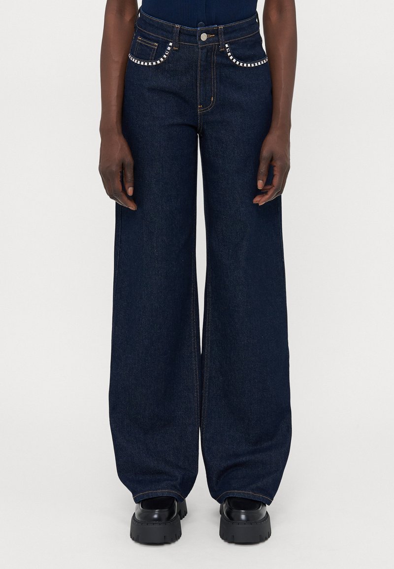 Vide leg mørke denimjeans med høy midje, med naglede detaljer på lommene og oransje søm, kombinert med kraftige svarte sko.