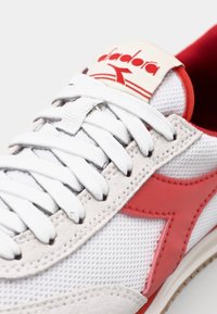 White mesh sneaker with red accents. Features a prominent red logo, flat white laces, and a textured beige overlay along the sides.