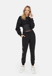 Felpa nera cropped con dettagli a stella rosa e coulisse, abbinata a jogger neri con disegni a stella rosa e polsini elasticizzati. Sneakers bianche.