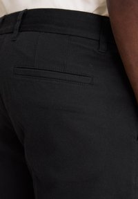 Czarne spodnie o gładkiej fakturze, z jedną tylnią kieszenią i subtelnym detalem brandingowym. Tkanina wydaje się wytrzymała i trwała.