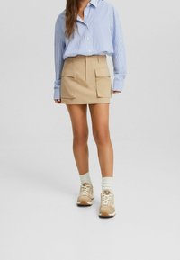 Kvinna iklädd en blå randig långärmad skjorta instoppad i en beige minikjol med stora framfickor och beige sneakers med vita strumpor.