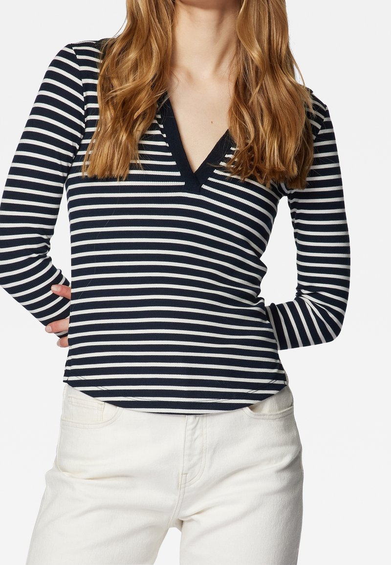 Námořnická a bílá pruhovaná halenka s dlouhým rukávem a výstřihem do V, zvlněným materiálem a mírně rozšířeným lemem, v kombinaci s bílými džínami s vysokým pasem.