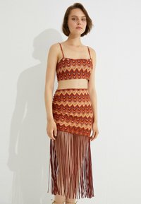 Zweiteiliges Outfit bestehend aus einem bauchfreien Oberteil und einem fransigen Rock. Beides in gemustertem Orange, Rot und Gold, mit dünnen Trägern und fließenden Fransen-Details.