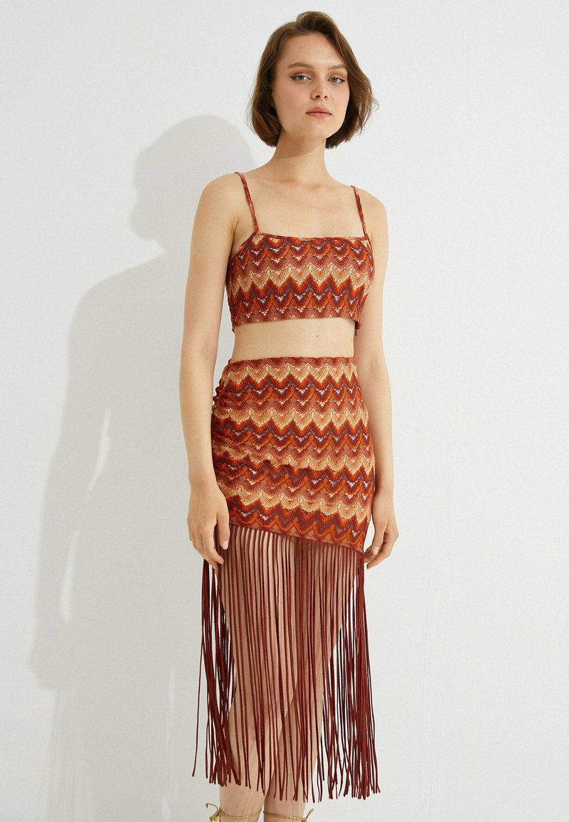 Sæt bestående af to dele med en cropped top og en frynse-nederdel. Begge er i mønstrede nuancer af orange, rød og guld, med tynde stropper og flydende frynse-detaljer.