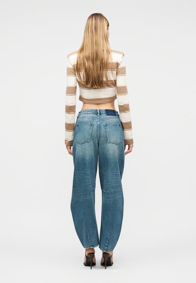 Γυναίκα με μακριά ξανθά μαλλιά που φοράει μια ριγέ μπεζ και λευκή μακρυμάνικη κοντή μπλούζα, φαρδιά τζιν παντελόνια και μαύρες γόβες, με την πλάτη στραμμένη.