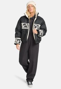 Puffjakke, sort med hvid bølgemønster, med en cremefarvet krave og manchetter. Parret med sorte overalls og sorte sneakers.
