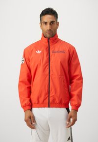 Röd nylonjacka med dragkedja framtill, elastiska muddar och midjeband. Har blå text "REVOLUTION" och en svart fotbollsgrafik på ärmen.