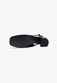 Sorte hæl-sko med en glat læderoverdel, firkantet tå og blokhæl. Har en minimalistic strap og en tekstureret sål.