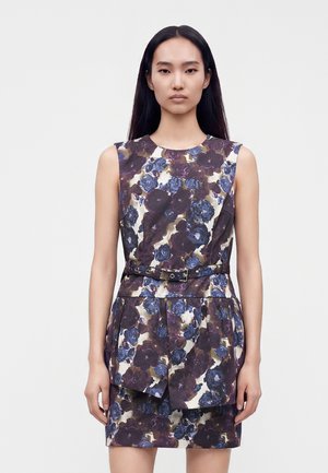 Jeune femme aux longs cheveux noirs portant une robe fleurie sans manches avec une ceinture, debout devant un fond gris clair uni.