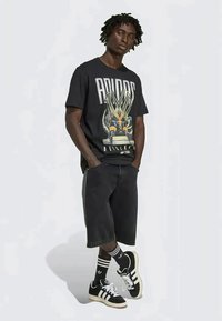 Vīrietis, ģērbies melnā Adidas grafiskā T-kreklā, melnās garās šortos, melnās Adidas zeķēs un melnās Adidas kedās, stāv ar rokām kabatās.