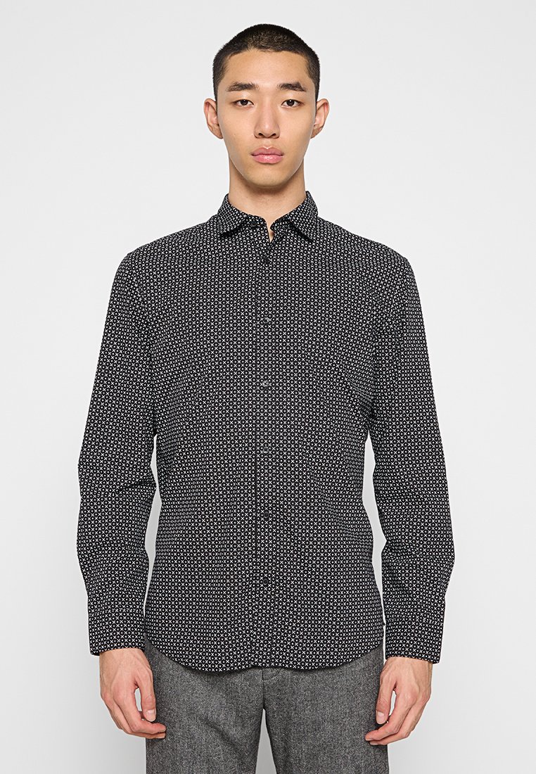 Man met een zwart overhemd met witte kruispatronen en een grijze broek, staande tegen een effen lichte achtergrond.