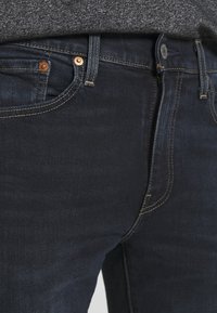 Mörkblå jeans med en slät textur, som har kopparnitar, gula sömmar och en klassisk femficksdesign.