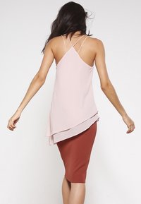 Vrouw in een lichtroze mouwloze top met kruislings banden en een gelaagde zoom, gecombineerd met een roestkleurige kokerrok, gezien van achteren.