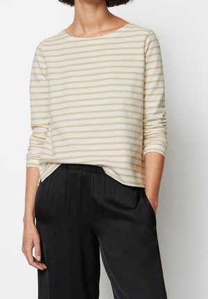 Persoon draagt een beige-wit gestreept shirt met lange mouwen, ingestopt in losse zwarte broek met elastische tailleband, staat met één hand in de zak.