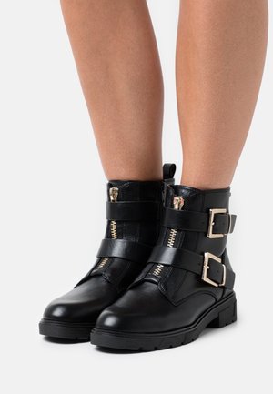 Černé kotníkové boty s texturovaným koženým povrchem, doplněné zlatými zipy, dvěma pásky s přezkami a nízkým, robustním podpatkem.