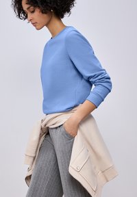 Femme aux cheveux bouclés portant un sweat-shirt bleu et un pantalon gris à fines rayures, une veste beige nouée autour de la taille, regardant vers le bas avec la main dans la poche.
