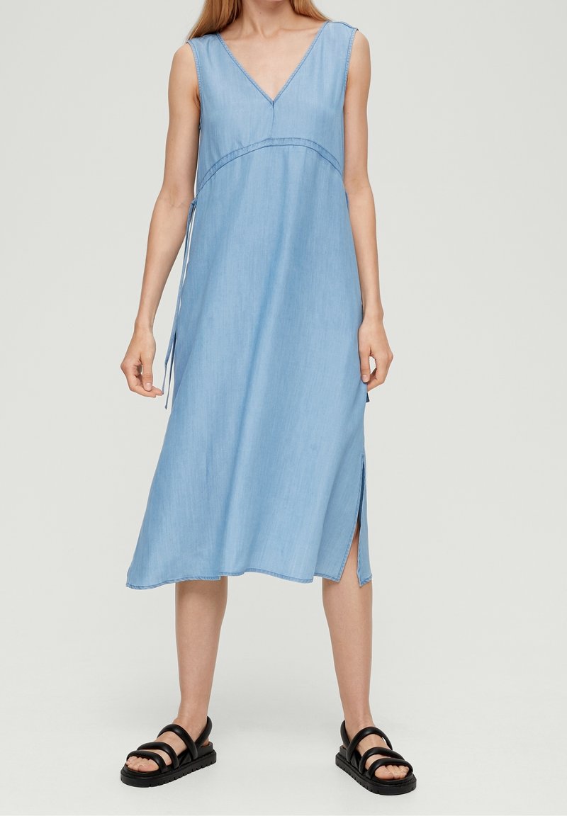 Femme portant une robe midi sans manches bleu clair à encolure en V avec fente latérale et sandales noires à brides, debout sur un fond uni.