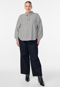 Naine seisab seljas lahtine must-valge vertikaalsete triipudega särk, laiade jalgadega mustad püksid ja studidega mustad chelsea saapad.