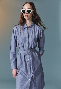 Stripet blå og hvit skjortekjole med lange ermer, belte i midjen, knappelukking og liten krage, kombinert med hvite geometriske solbriller.
