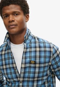 Camicia di flanella a quadri blu e neri con accenti beige, dotata di colletto button-down e una patch con logo sul petto. Maglietta bianca visibile.