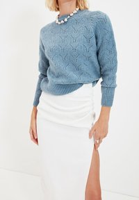 Hellblauer Strickpullover mit strukturiertem, gemustertem Design und gerippten Kanten, kombiniert mit einem weißen Wickelrock mit einem seitlichen Schlitz.