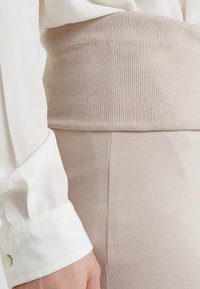 Beige žebrovaný pás s texturovaným povrchem, spojený s hladkou béžovou tužkovou sukní a bílou košili s knoflíkovými manžetami.