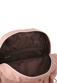 Interior de mochila de tela suave en color rosa con un forro marrón, un bolsillo con cremallera y el logo de DKNY visible en el interior. Textura plana y suave.