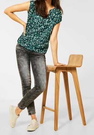 Grønne mønstrede, kortærmede toppar parret med grå tætsiddende jeans. Modellen læner sig op ad en lys træstol med et enkelt design. Hvide sneakers.
