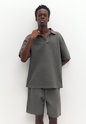 Junger Mann mit kurzen lockigen Haaren, der ein lockeres graues Poloshirt und passende Shorts trägt, steht vor weißem Hintergrund mit erhobenem Arm.