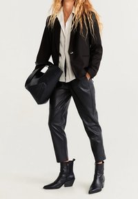 Blazer nero su misura sopra una camicia bianca con bottoni, abbinato a pantaloni in ecopelle nera e stivaletti; porta una borsa nera.