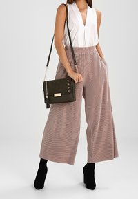 Hellrosa gestreifte, weite Hosen mit hohem Bund, kombiniert mit einer weißen, ärmellosen Bluse und einer dunkelgrünen Umhängetasche. Schwarze Stilettos.