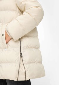 Dunjakke i lys beige med quiltet design. Har en lynlåslomme og knaplukket klap. Blød tekstur med et tykt, isoleret udseende.