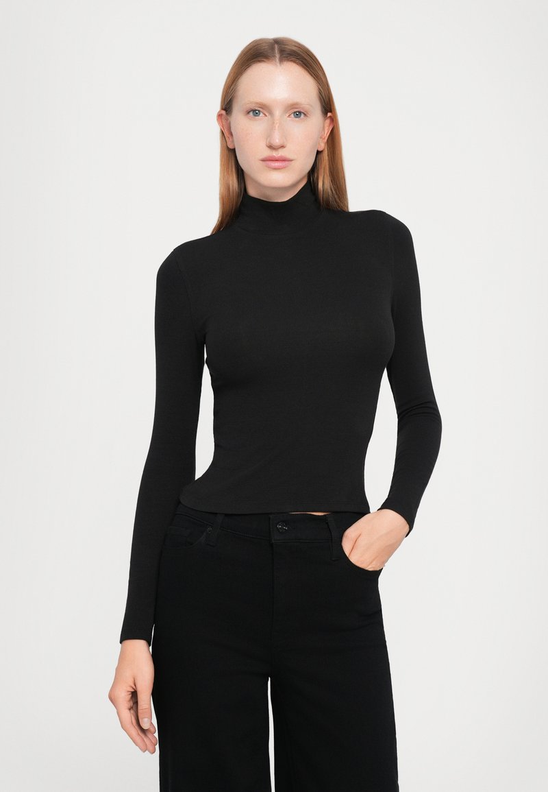 Femme avec des cheveux raides rouges portant un col roulé noir ajusté et un pantalon noir, se tenant avec une main dans sa poche devant un fond blanc.