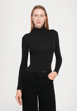 Kvinne med rett rødt hår iført en tettsittende svart turtleneck og svarte bukser, stående mot en ensfarget lys bakgrunn.