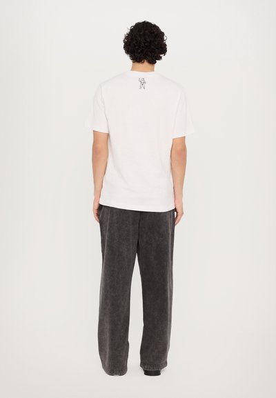 T-shirt en coton blanc à manches courtes, avec un petit graphique d'astronaute dans le dos. Porté avec un pantalon ample de couleur gris foncé.