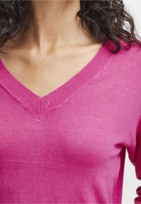 Pull à col en V de couleur rose vif, fabriqué en tissu tricoté doux. Présente des bordures côtelées au niveau de l'encolure. Texture lisse, légère élasticité, coupe ajustée.