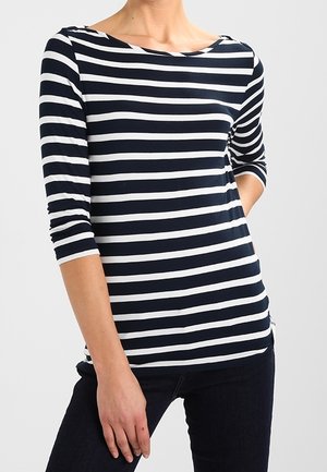 Vrouw die een zwart-wit horizontaal gestreept top met driekwart mouwen en een donkere broek draagt, staand met één hand in de zak.