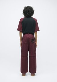 Persoon met krullend haar, gekleed in een zwart en bordeaux rood kort mouwtop en bijpassende wijde broek met een boompje logo op de achterzak.