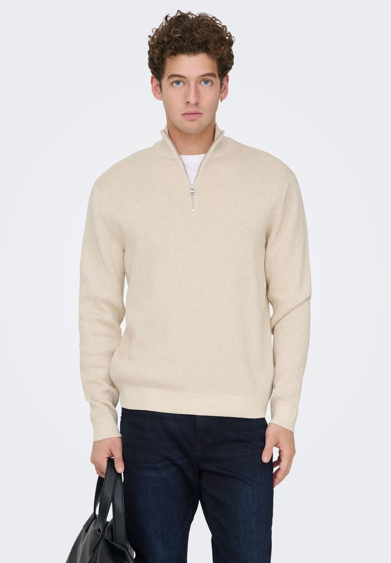 Beige zip-up sweater with a soft texture and long sleeves, paired with dark jeans. Model holds a black bag, standing against a light background.