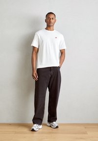 Hvid bomulds t-shirt med rødt logo, parret med sorte løse bukser og grå sportssko, stående på træbelægning mod en lys væg.