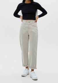Femme portant un pantalon beige taille haute, un crop top noir à manches longues et des baskets blanches, debout les mains sur les hanches.