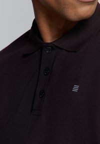 Zwarte polo met een gestructureerde stof, een drie-knopen placket en een klein grijs gestreept logo op de linkerborst.