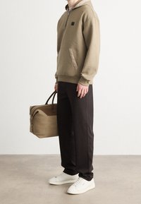 Khaki bluza z kapturem na suwak i z przednią kieszenią, w połączeniu z czarnymi spodniami i białymi tenisówkami. Trzyma beżowo-brązową, usztywnianą torebkę.