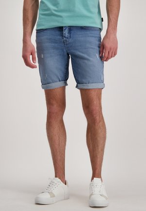 Lichtblauwe denimshorts met opgerolde manchetten en versleten details, gecombineerd met witte sneakers. De stof heeft een gladde textuur.