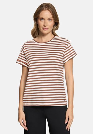 Woman with light brown hair wearing a white and brown horizontal striped short-sleeve shirt and black pants, standing against a plain background.