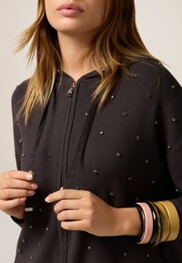 Haut de sweat à capuche noir zippé avec de petits strass, cordons au niveau du col, et différents bracelets colorés au poignet. Texture de tissu lisse.