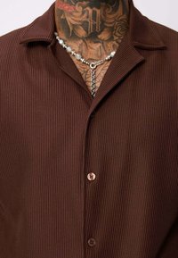 Chemise marron à manches courtes avec une texture côtelée, dotée d'un col pointu et d'une fermeture à un bouton. Accentuée par un collier en argent.