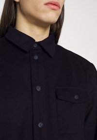 Dark navy button-up shirt with a collar, featuring a left chest pocket, and smooth texture. Buttons are also navy in color.
